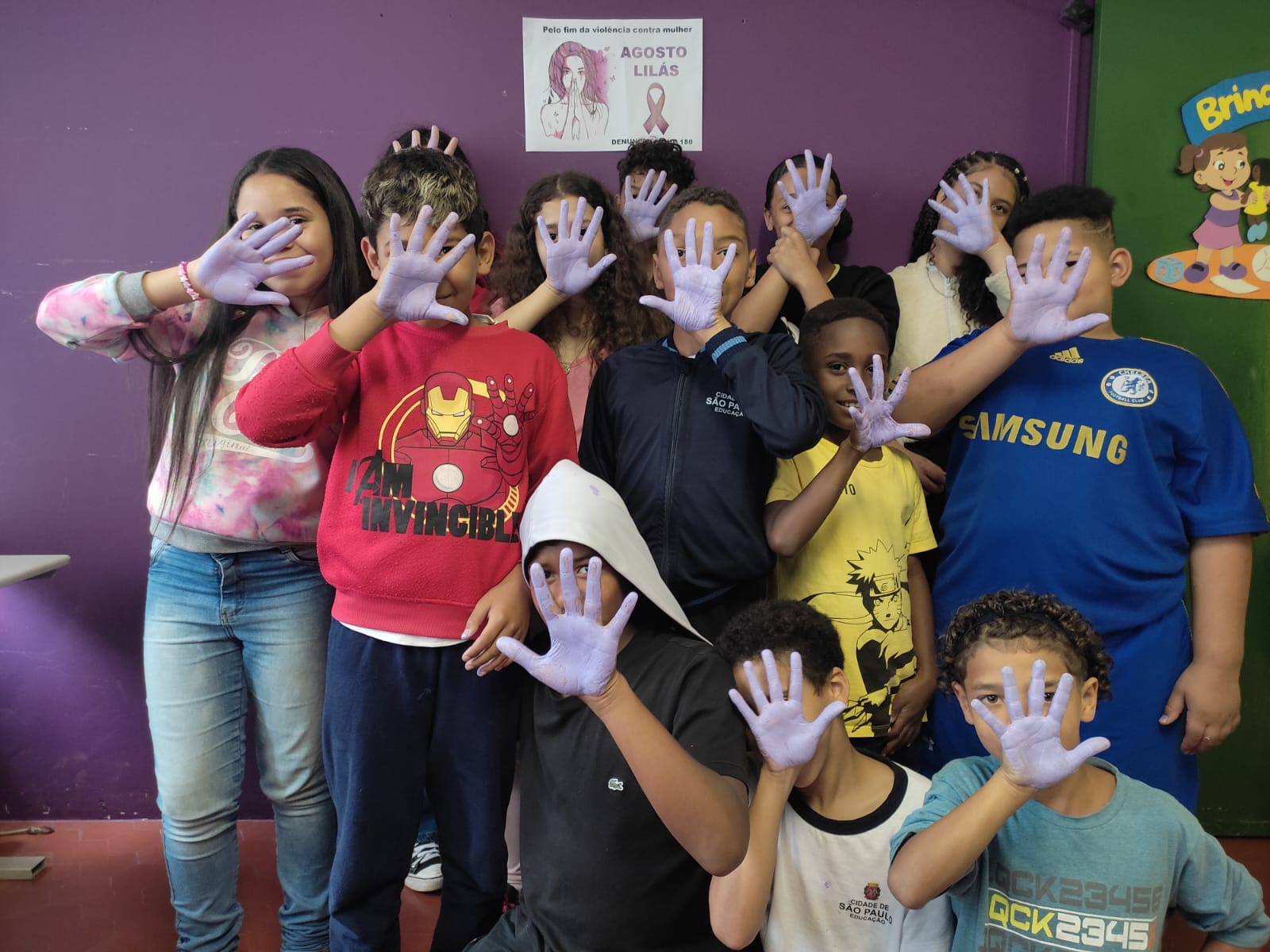 Na imagem, outras crianças em pé com as mãos pintadas, enquanto outras três estão agachadas, cobrindo o rosto com as mãos